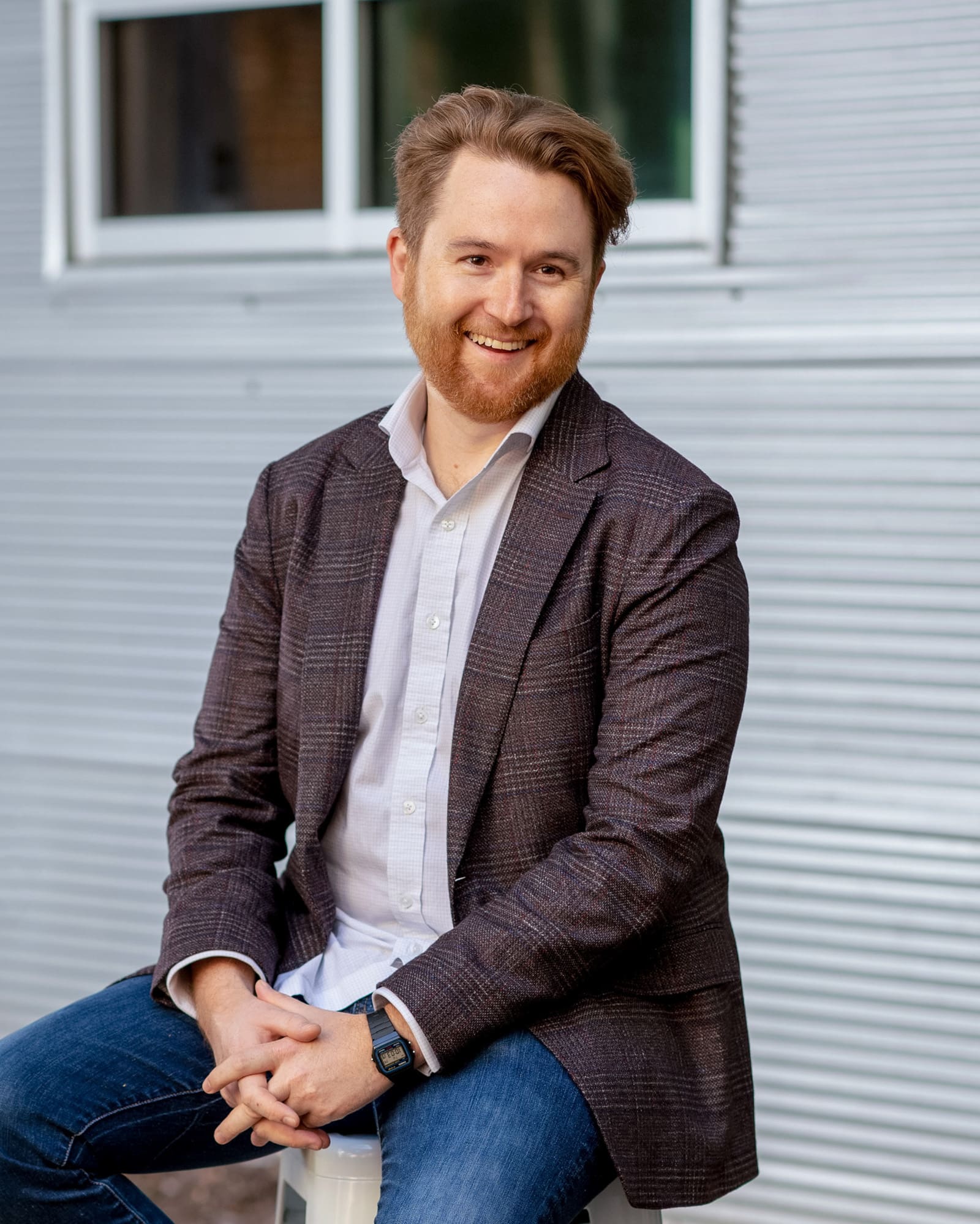 Jason Leiss smiles sitting on a chair in front of an aluminum wall and window.